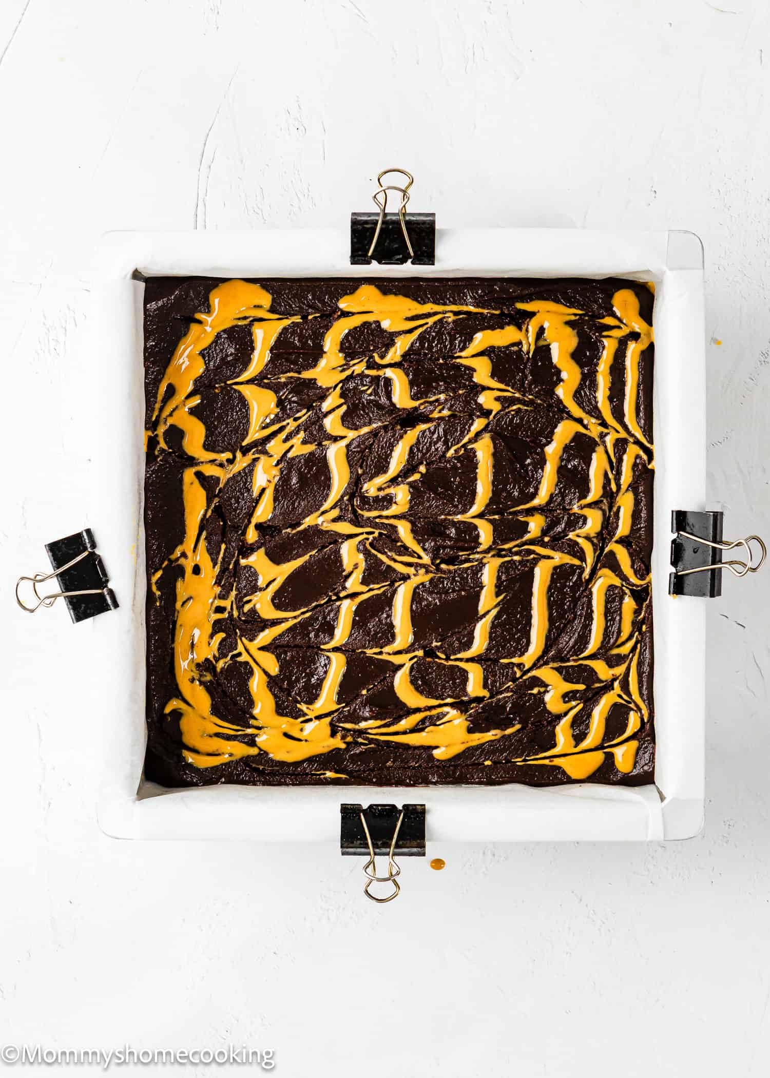 A square baking pan lined with parchment paper and binder clips holds chocolate batter swirled with peanut butter on top, ready to be baked.