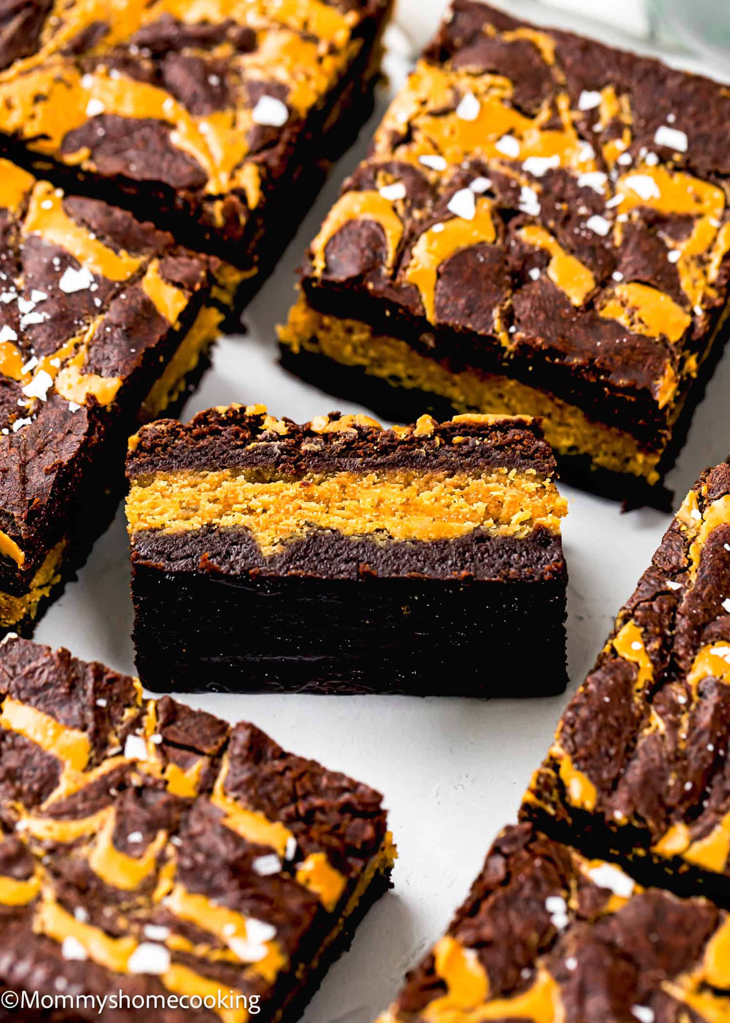 Close-up of layered brownie bars with a chocolate and peanut butter swirl, topped with sea salt flakes, arranged on a white surface.