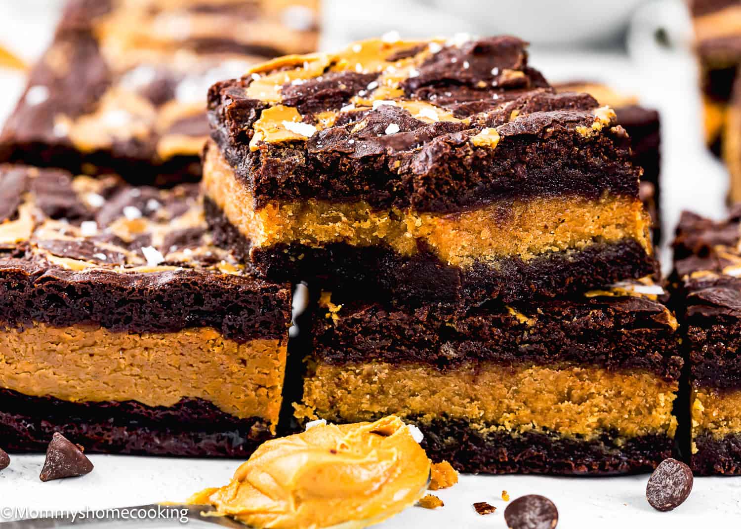 Eggless Peanut Butter Brownies with a thick layer of creamy filling, topped with a drizzle of chocolate and a sprinkle of sea salt, displayed on a white surface.