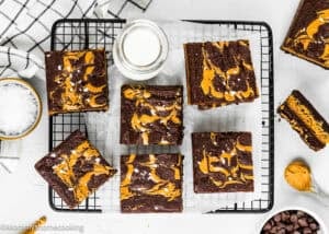 Six eggless peanut butter brownies sit on a wire rack with parchment paper, surrounded by a small bowl of salt, a jar of milk, and chocolate chips.