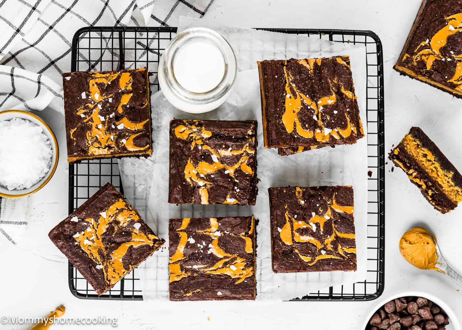 Six eggless peanut butter brownies sit on a wire rack with parchment paper, surrounded by a small bowl of salt, a jar of milk, and chocolate chips.