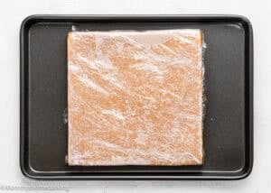 A square block of dough wrapped in plastic wrap sits on a black baking tray.