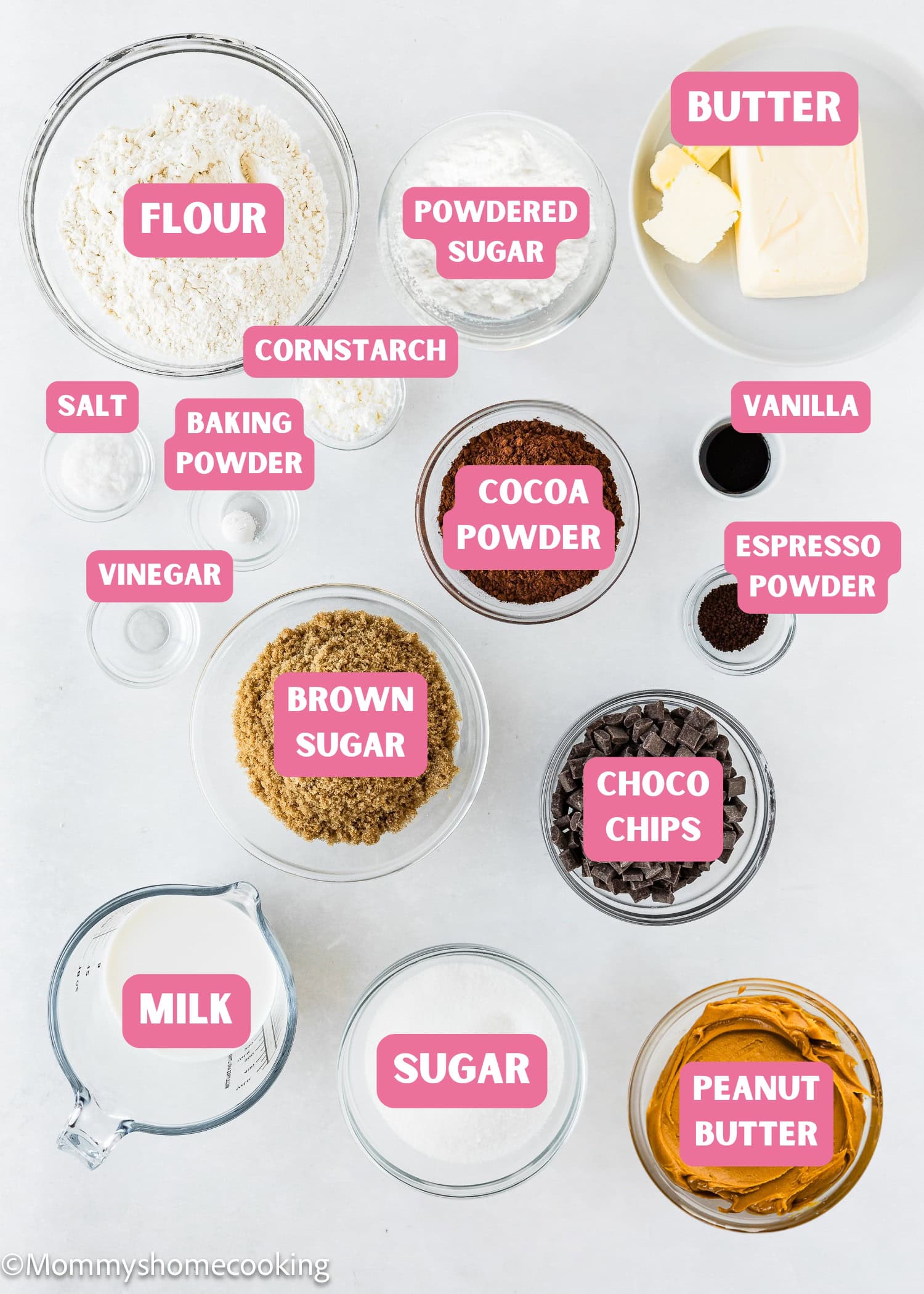 Top-down view of labeled Eggless Peanut Butter Brownies ingredients—flour, sugar, cocoa powder, peanut butter, milk, butter, and more—in bowls and containers on a white surface.