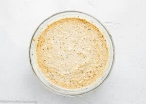 A glass bowl filled with a creamy, seasoned mixture of Eggless Protein Baked Oatmeal, featuring visible herbs and small chunks, sits on a white textured surface.