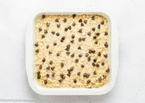 A square white baking dish filled with unbaked Eggless Protein Baked Oatmeal, topped with scattered chocolate chips, sits on a light surface.