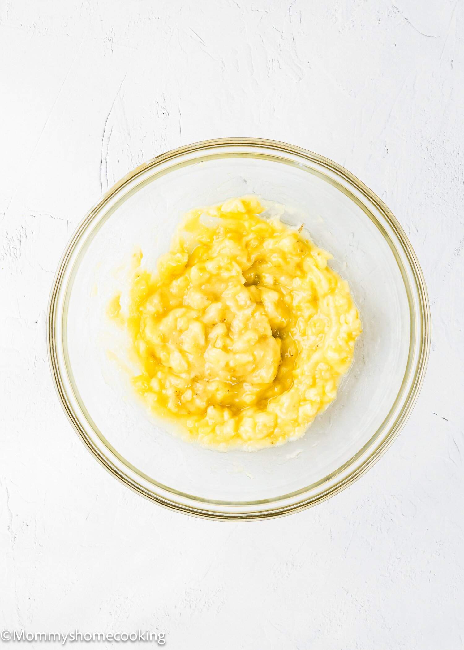 A glass bowl filled with mashed bananas sits on a white textured surface, showing a yellow, slightly thicker mixture.