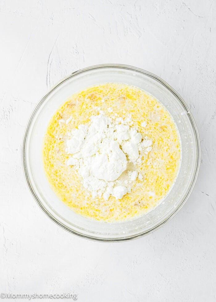 A glass bowl filled with a mixture of wet ingredients and a mound of white cottage cheese on top, set on a white textured surface.