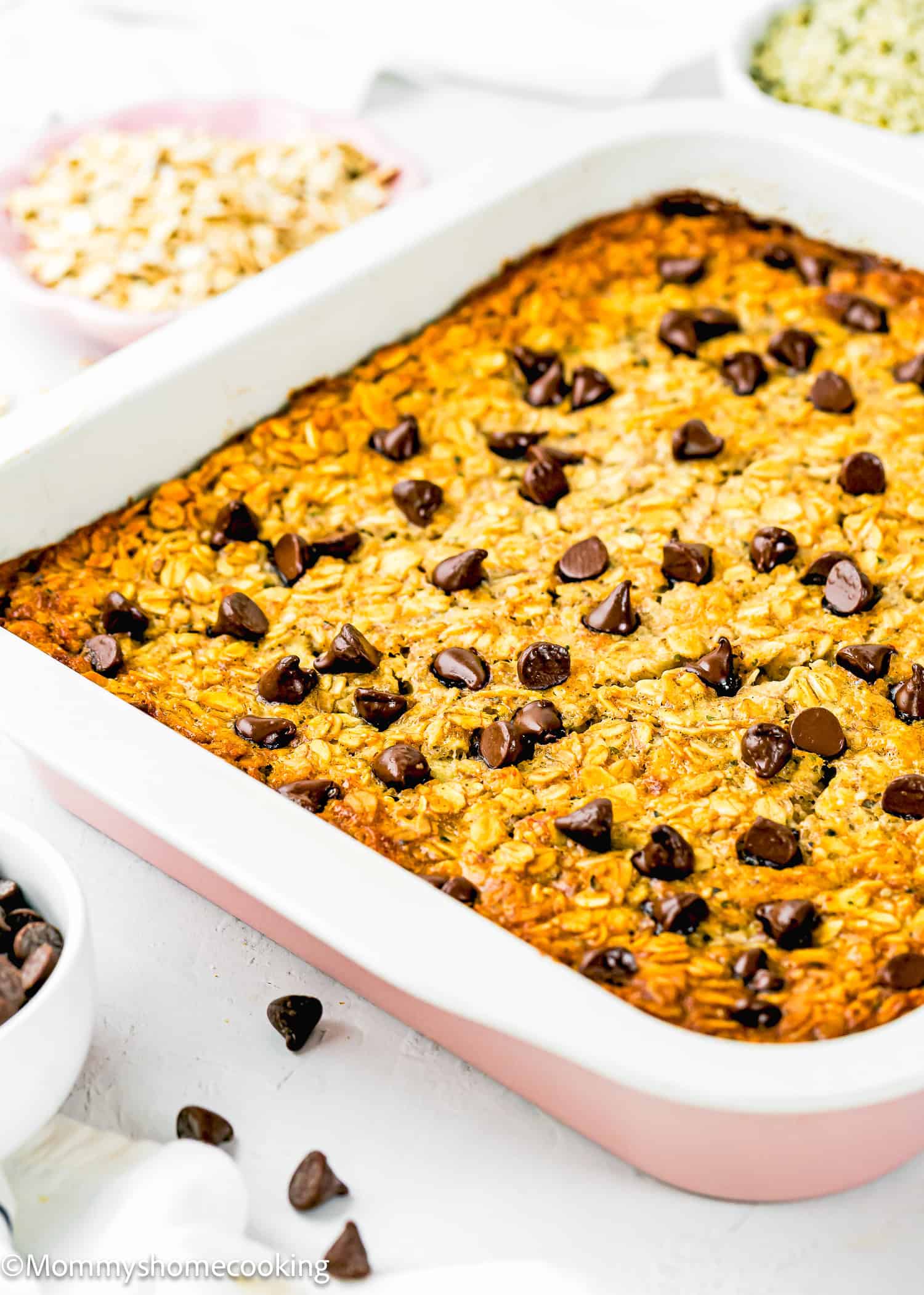 An Eggless Protein Baked Oatmeal casserole topped with chocolate chips sits in a pink-bordered baking dish, surrounded by small bowls of oats and chocolate chips on a white surface.