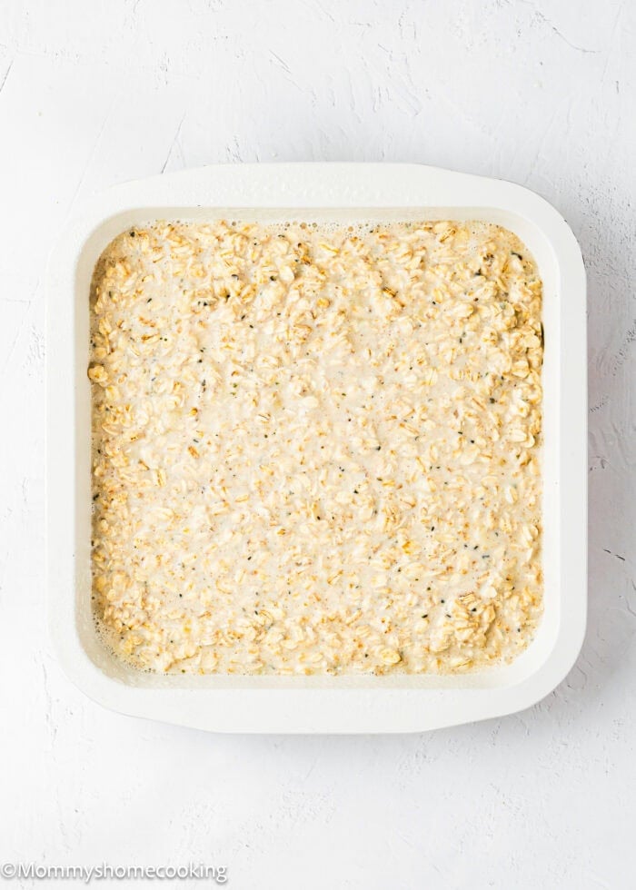 A white square baking dish filled with a baked mixture of oats and liquids sits on a freshly baked surface.