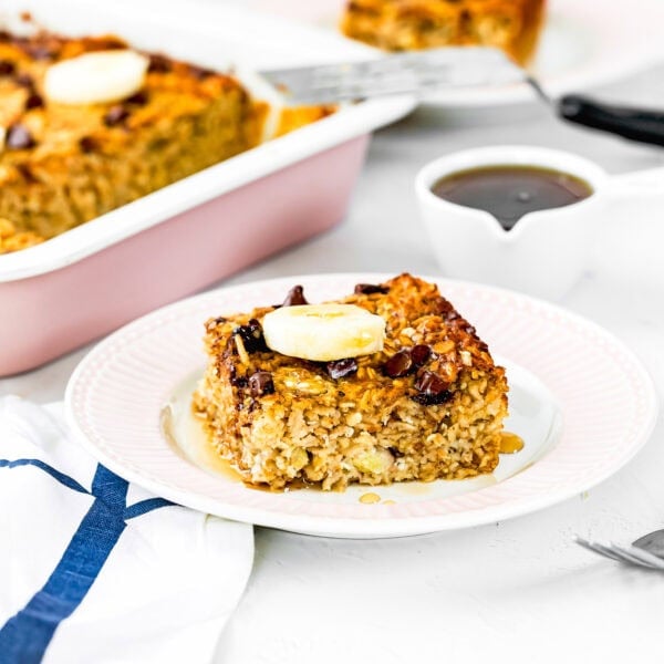 A square slice of Eggless Protein Baked Oatmeal topped with banana and syrup sits on a white plate. More baked oatmeal is in a pink baking dish in the background, with a spatula and small pitcher of syrup nearby.