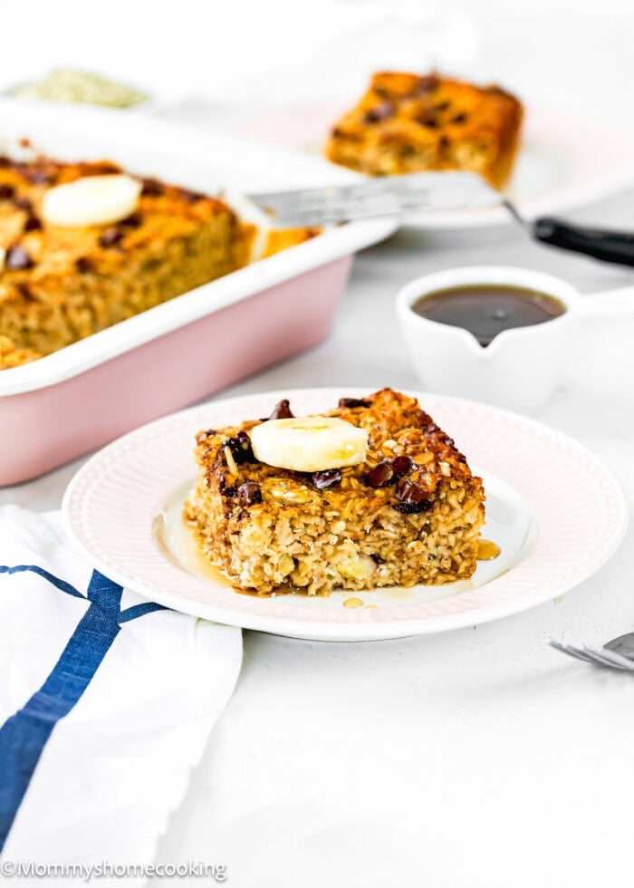 A square slice of Eggless Protein Baked Oatmeal topped with banana and syrup sits on a white plate. More baked oatmeal is in a pink baking dish in the background, with a spatula and small pitcher of syrup nearby.