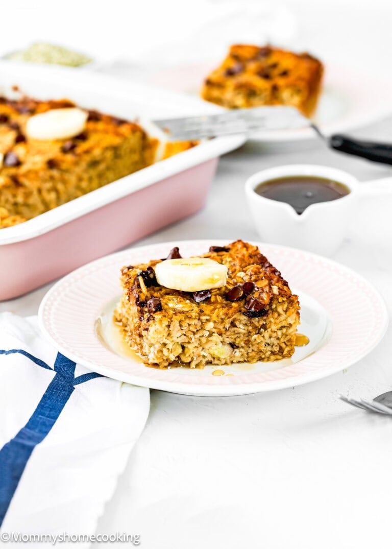 A square slice of Eggless Protein Baked Oatmeal topped with banana and syrup sits on a white plate. More baked oatmeal is in a pink baking dish in the background, with a spatula and small pitcher of syrup nearby.