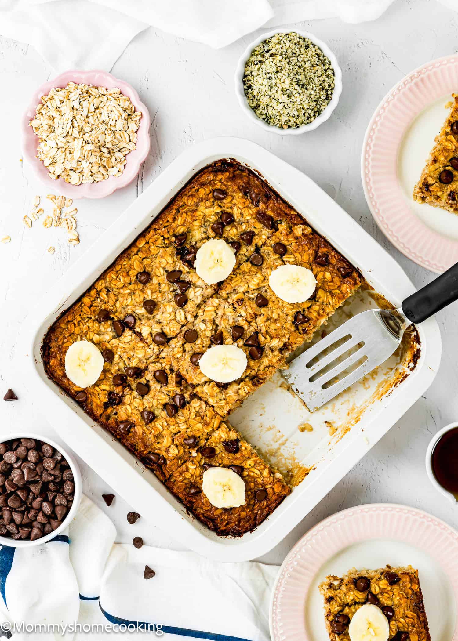 Lift a spatula in one piece, a square baking dish filled with egg-free protein-baked oatmeal topped with banana slices and chocolate chips. The oats, chocolate chips and seeds are located in a small bowl nearby. The oatmeal plate is sitting on the side.