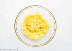A glass bowl filled with mashed bananas, perfect for preparing Eggless Protein Baked Oatmeal, placed on a white textured surface.