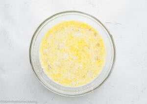 A glass bowl filled with a yellow, bubbly Eggless Protein Baked Oatmeal mixture sits on a white textured surface.