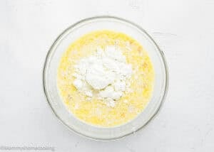 A glass bowl filled with a yellow liquid mixture and a mound of flour sits on a white textured surface, ready to be mixed for Eggless Protein Baked Oatmeal.