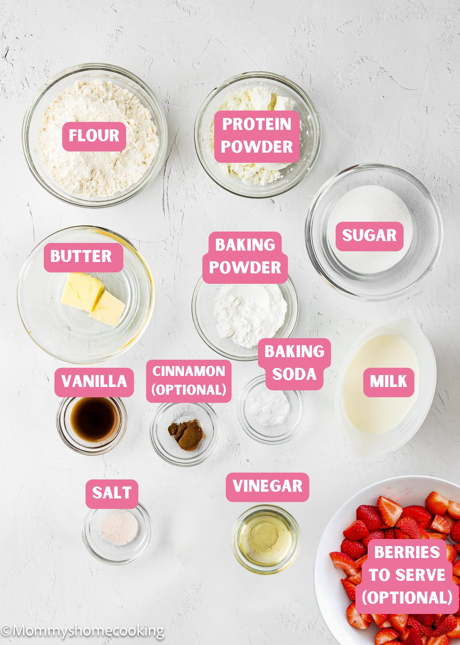 A flat lay of labeled baking ingredients in bowls for Eggless Protein Pancakes (Fluffy & Tasty): flour, protein powder, sugar, baking powder, baking soda, milk, butter, vanilla, cinnamon, salt, vinegar, and optional berries.
