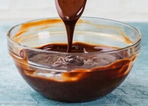 A spoon drizzles Foolproof Chocolate Ganache (With Dairy-Free Option!) into a glass bowl filled with melted chocolate on a blue surface.