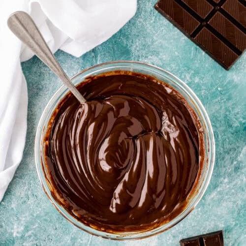 A glass bowl filled with smooth, melted Foolproof Chocolate Ganache (With Dairy-Free Option!) and a spoon inside sits next to pieces of chocolate on a blue textured surface.