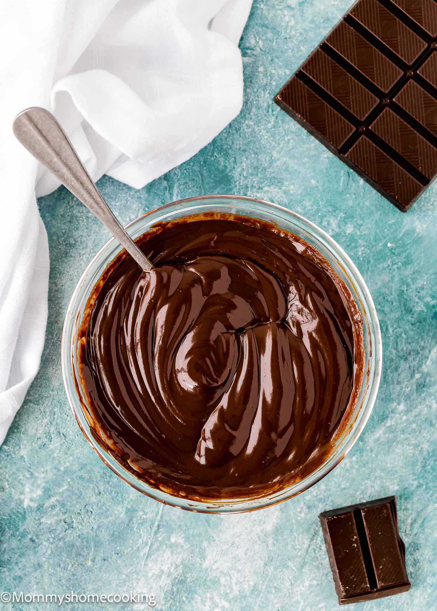 A glass bowl filled with smooth, melted Foolproof Chocolate Ganache (With Dairy-Free Option!) and a spoon inside sits next to pieces of chocolate on a blue textured surface.
