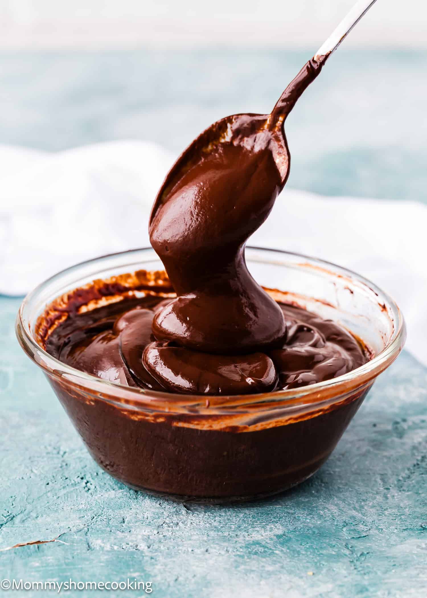 A spoon lifts thick, glossy Foolproof Chocolate Ganache (With Dairy-Free Option!) from a clear glass bowl set on a blue surface.