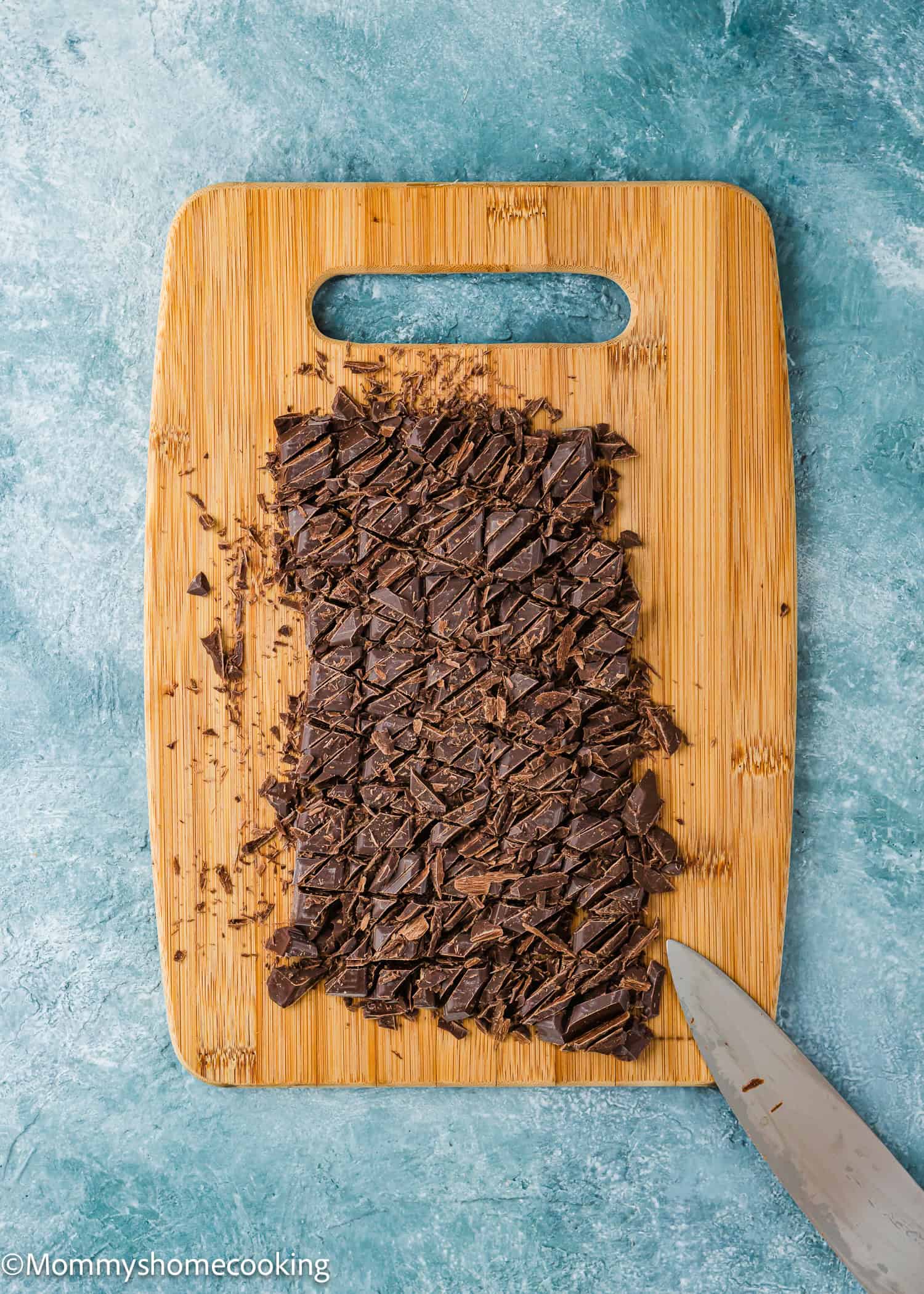 Chopped dark chocolate on a wooden cutting board with a knife on the side, ready to make foolproof chocolate ganache (with dairy-free option!)—all set on a blue textured surface.