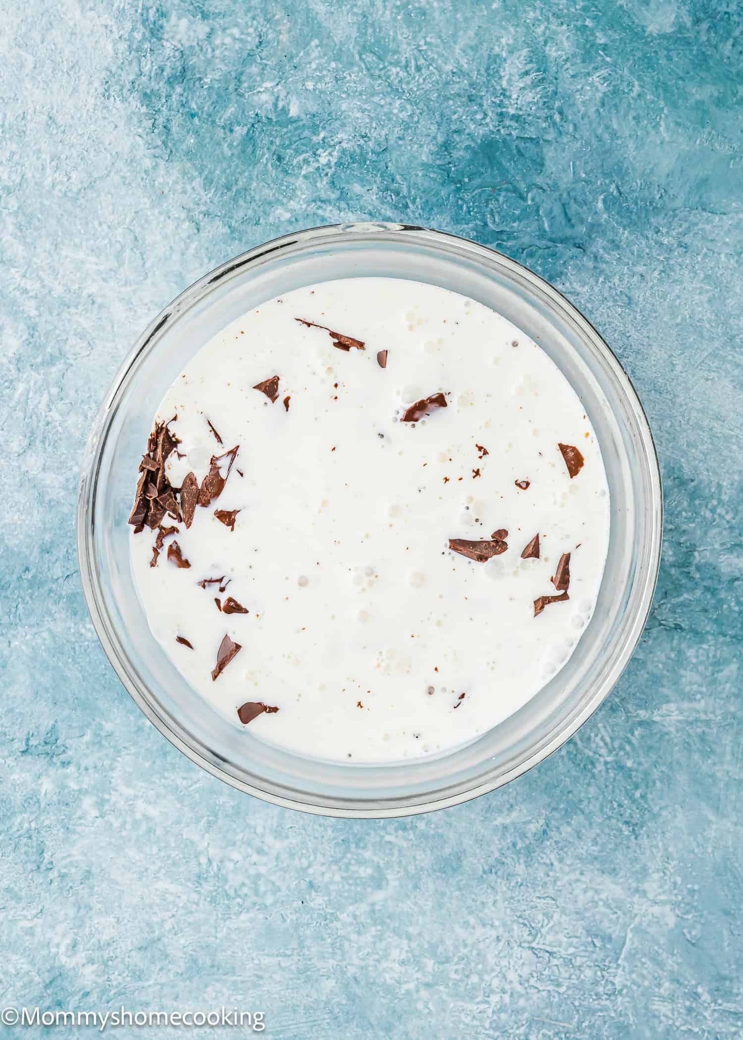 A glass bowl filled with white liquid, possibly whipped cream or mousse, topped with chocolate shavings—perfect as a base or topping for foolproof chocolate ganache (with dairy-free option!)—set on a blue textured surface.