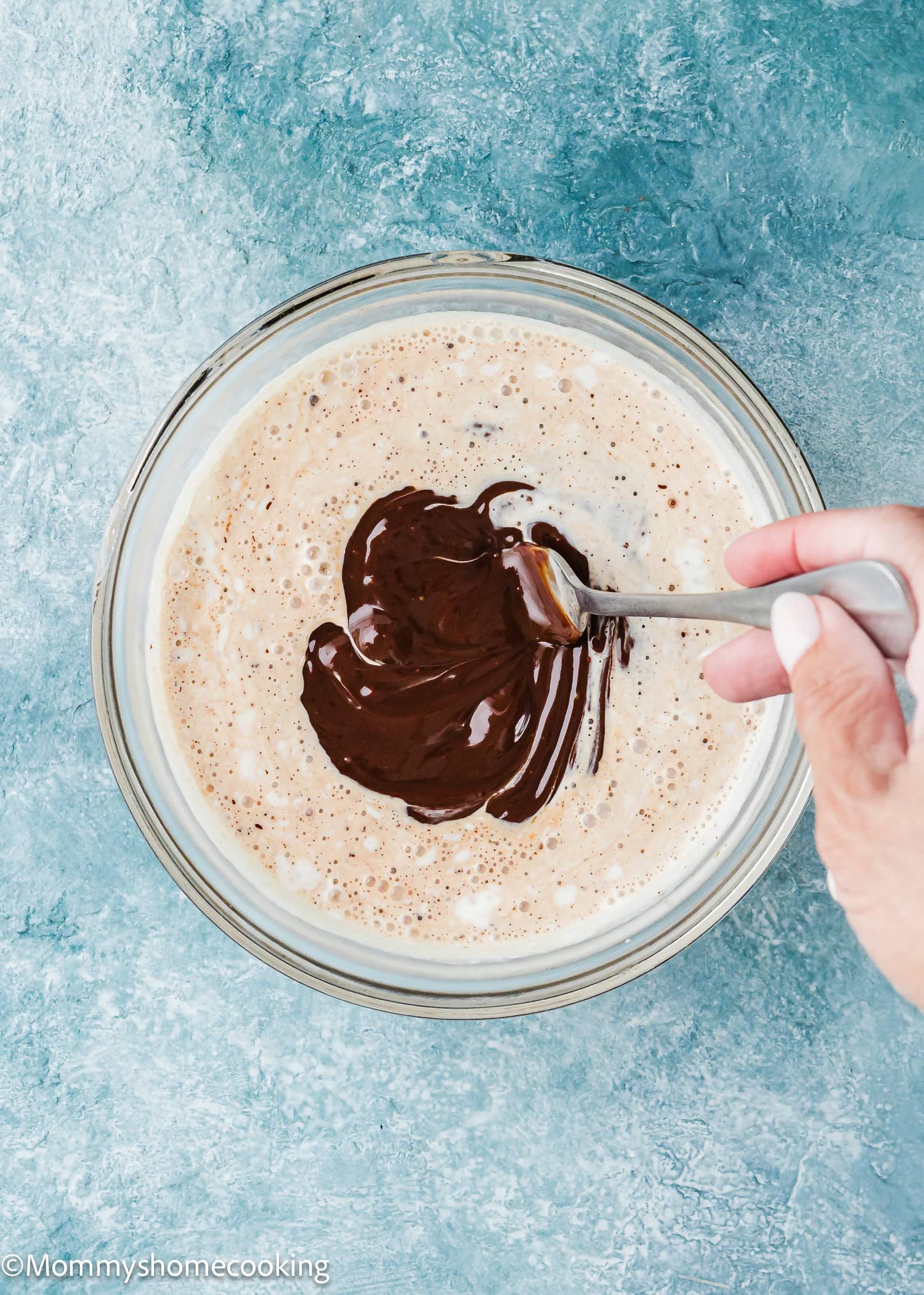 A hand holds a spoon, stirring chocolate and cream over a blue textured surface—just one step toward foolproof chocolate ganache (with dairy-free option!).