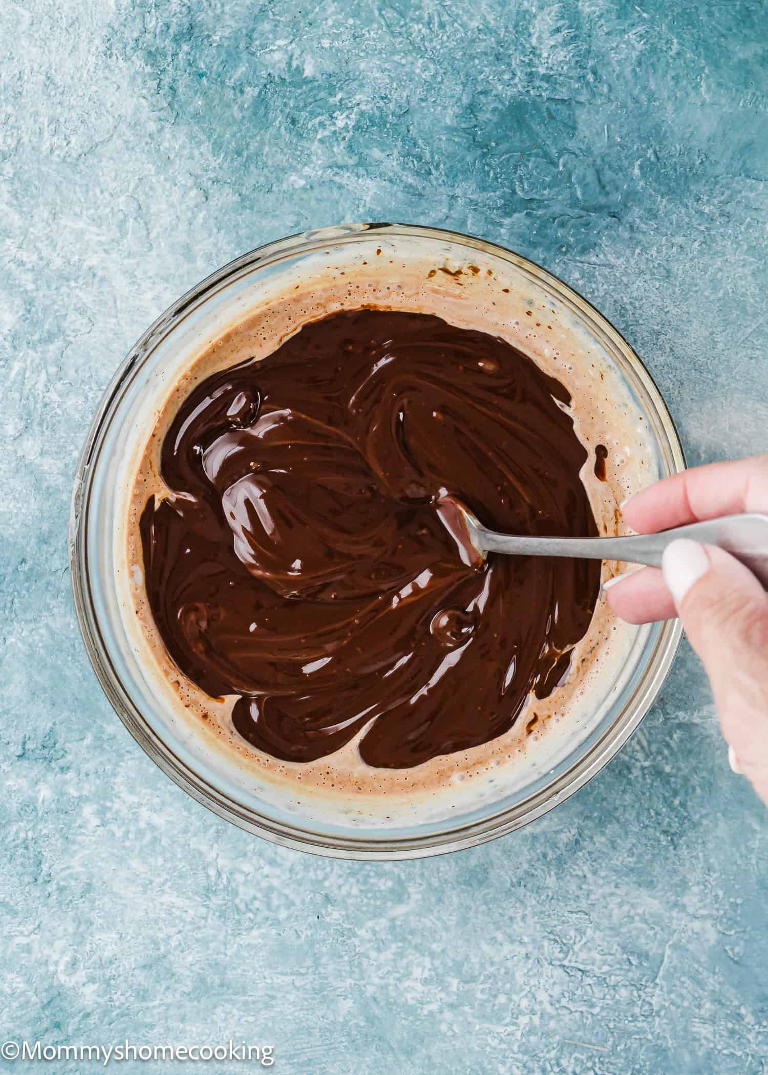 A hand stirs melted chocolate in a glass bowl using a spoon against a blue textured background, creating a foolproof chocolate ganache (with dairy-free option!) for any dessert occasion.