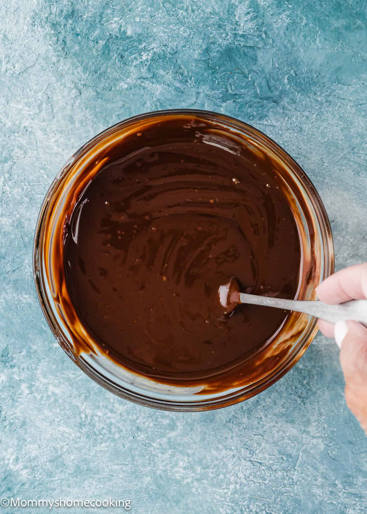 A hand stirs smooth, melted Foolproof Chocolate Ganache (With Dairy-Free Option!) in a glass bowl with a metal spoon, set on a blue textured surface.