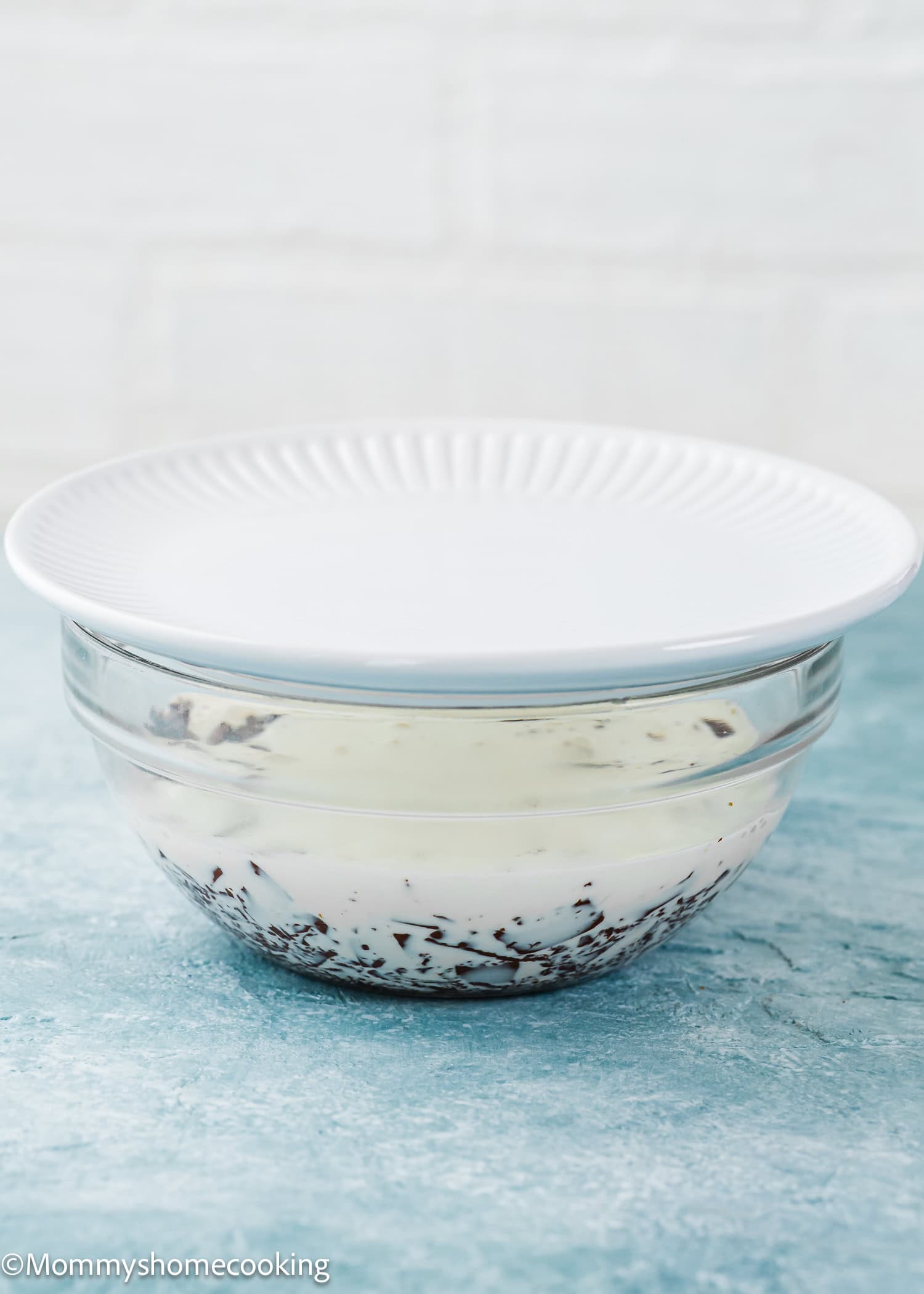 A glass bowl with a white plate on top, containing chocolate and cream, sits on a blue surface—perfect as a base for your foolproof chocolate ganache (with dairy-free option!).