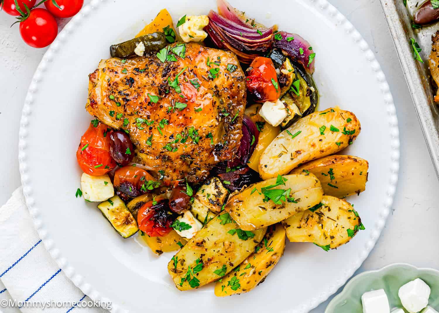 A white plate with roasted Greek chicken, Slow Cooker Lemon Greek Potatoes, zucchini, cherry tomatoes, red onion, olives, and feta cheese, all garnished with chopped parsley.