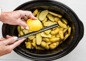 A person grates a lemon over sliced potatoes in a black slow cooker, preparing delicious Slow Cooker Lemon Greek Potatoes.