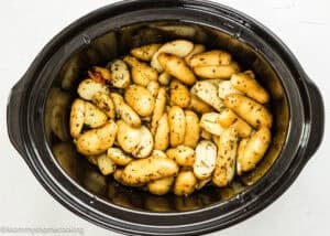 Sliced fingerling potatoes seasoned with herbs are shown cooking in a black slow cooker on a white surface, making delicious Slow Cooker Lemon Greek Potatoes.