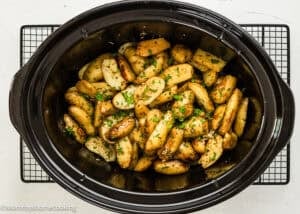 Slow Cooker Lemon Greek Potatoes: Sliced roasted potatoes with herbs and zesty lemon flavor in a black slow cooker, placed on a cooling rack.