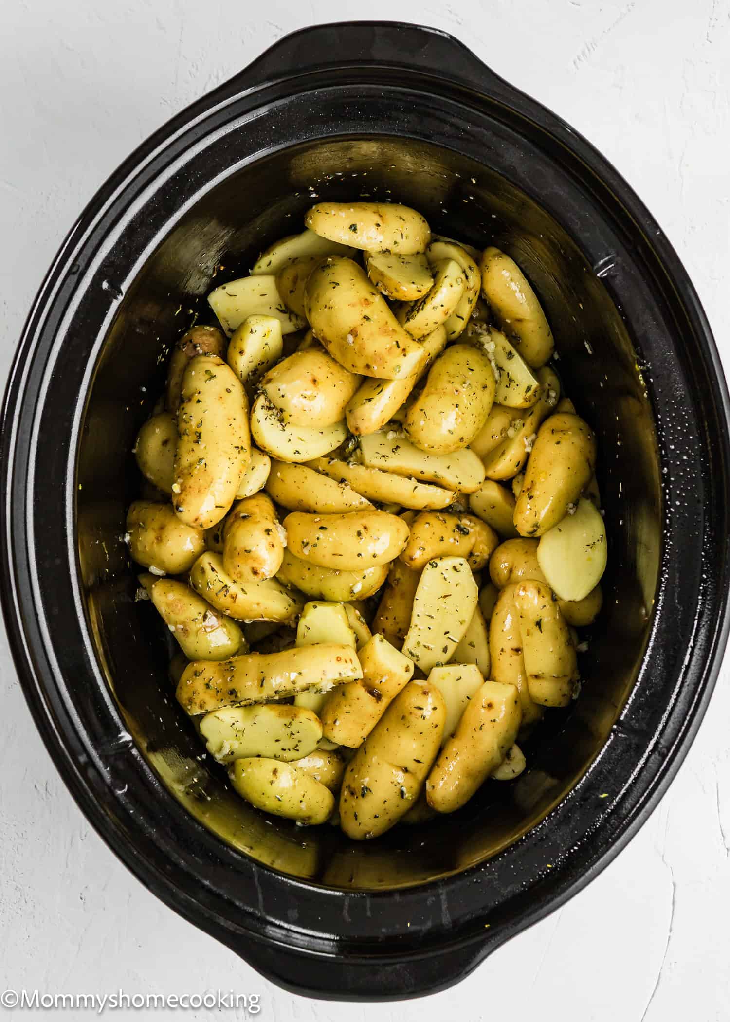 Fingerling potatoes seasoned with herbs and oil are placed inside a black slow cooker, ready to become delicious Slow Cooker Lemon Greek Potatoes.