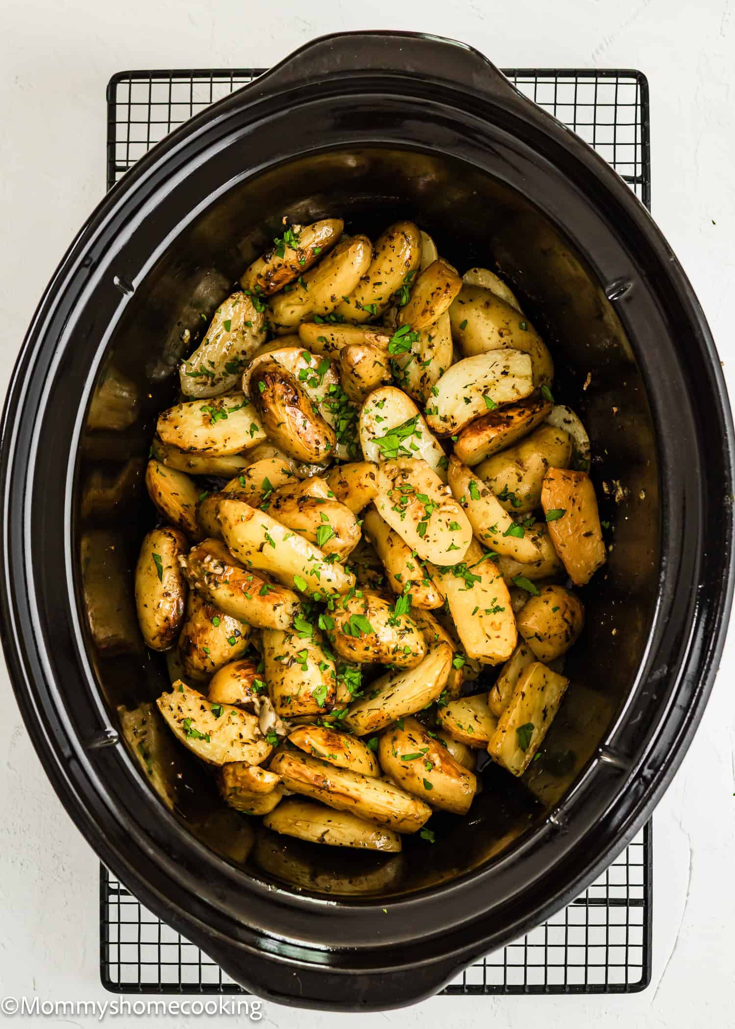 Slow Cooker Lemon Greek Potatoes: a slow cooker filled with sliced roasted potatoes, seasoned with herbs and lemon, then garnished with chopped parsley and placed on a cooling rack.