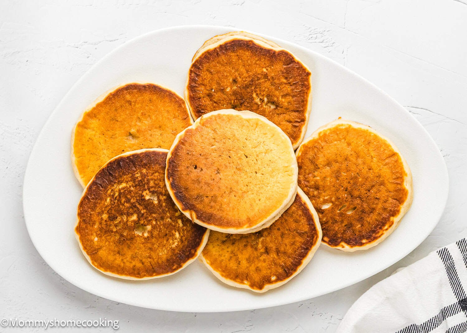 A white oval plate with seven golden-brown Eggless Protein Pancakes (Fluffy & Tasty) arranged in a circular pattern on a white surface.
