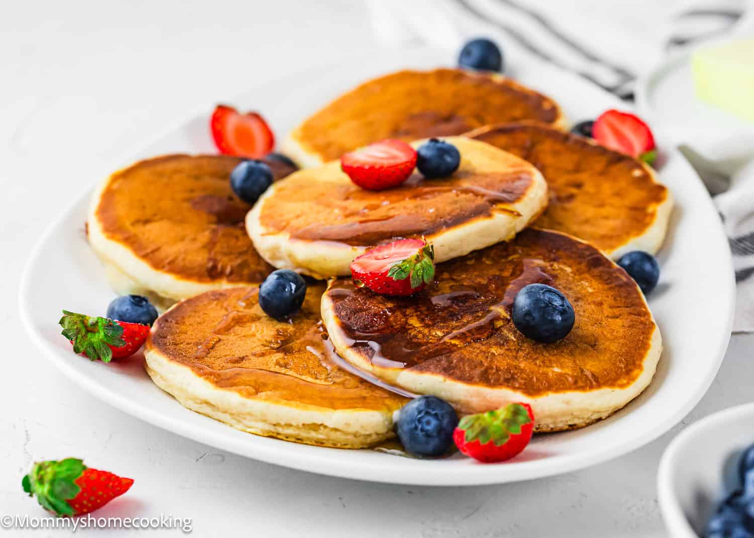 A white plate with five golden-brown Eggless Protein Pancakes (Fluffy & Tasty) topped with syrup, fresh blueberries, and sliced strawberries on a light background.