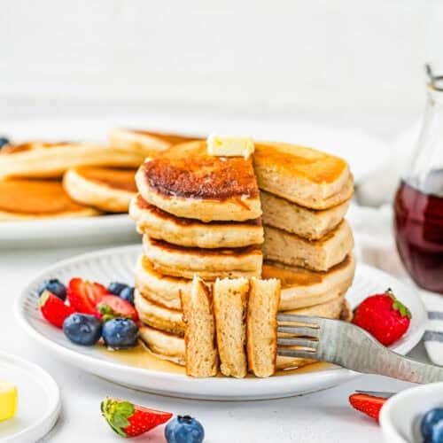 A stack of Eggless Protein Pancakes (Fluffy & Tasty) topped with butter is served with blueberries and strawberries, with a fork holding a sliced piece. Syrup and more pancakes are visible in the background.
