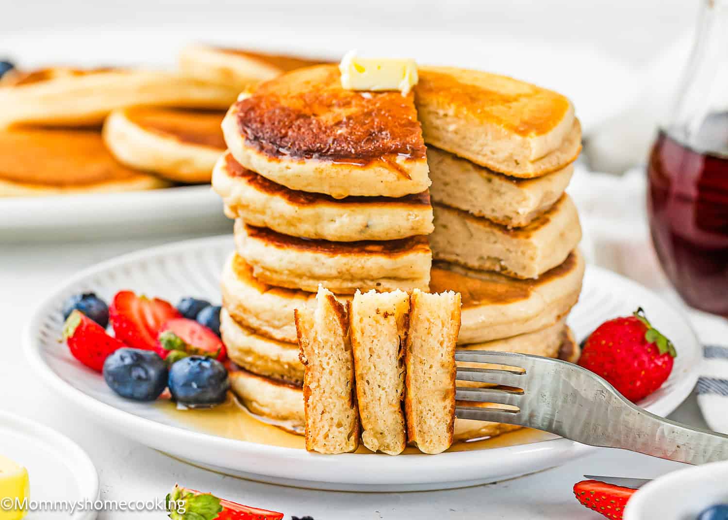 A stack of Eggless Protein Pancakes (Fluffy & Tasty) with a pat of butter, cut to reveal their soft inside, served with syrup, strawberries, and blueberries on a white plate.
