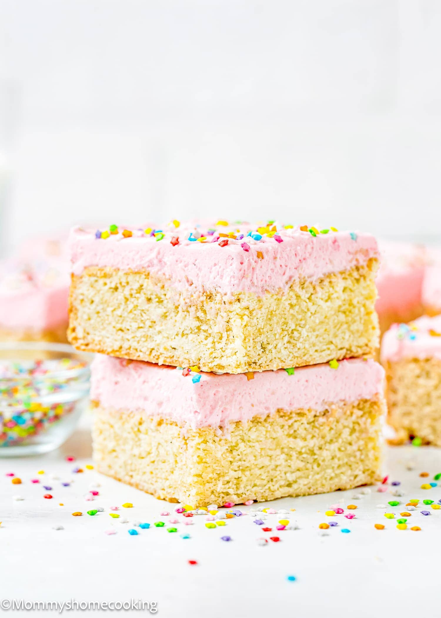 Two square vanilla cake bars with pink frosting and colorful sprinkles are stacked on a white surface—just one of 10 Delicious Egg-Free Bar Recipes You’ll Love, with more bars and a bowl of sprinkles in the background.
