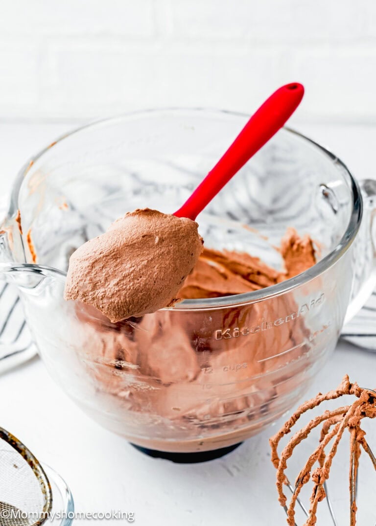 A glass mixing bowl with chocolate whipped cream and a red spatula resting on the rim; a whisk with chocolate remnants is nearby.