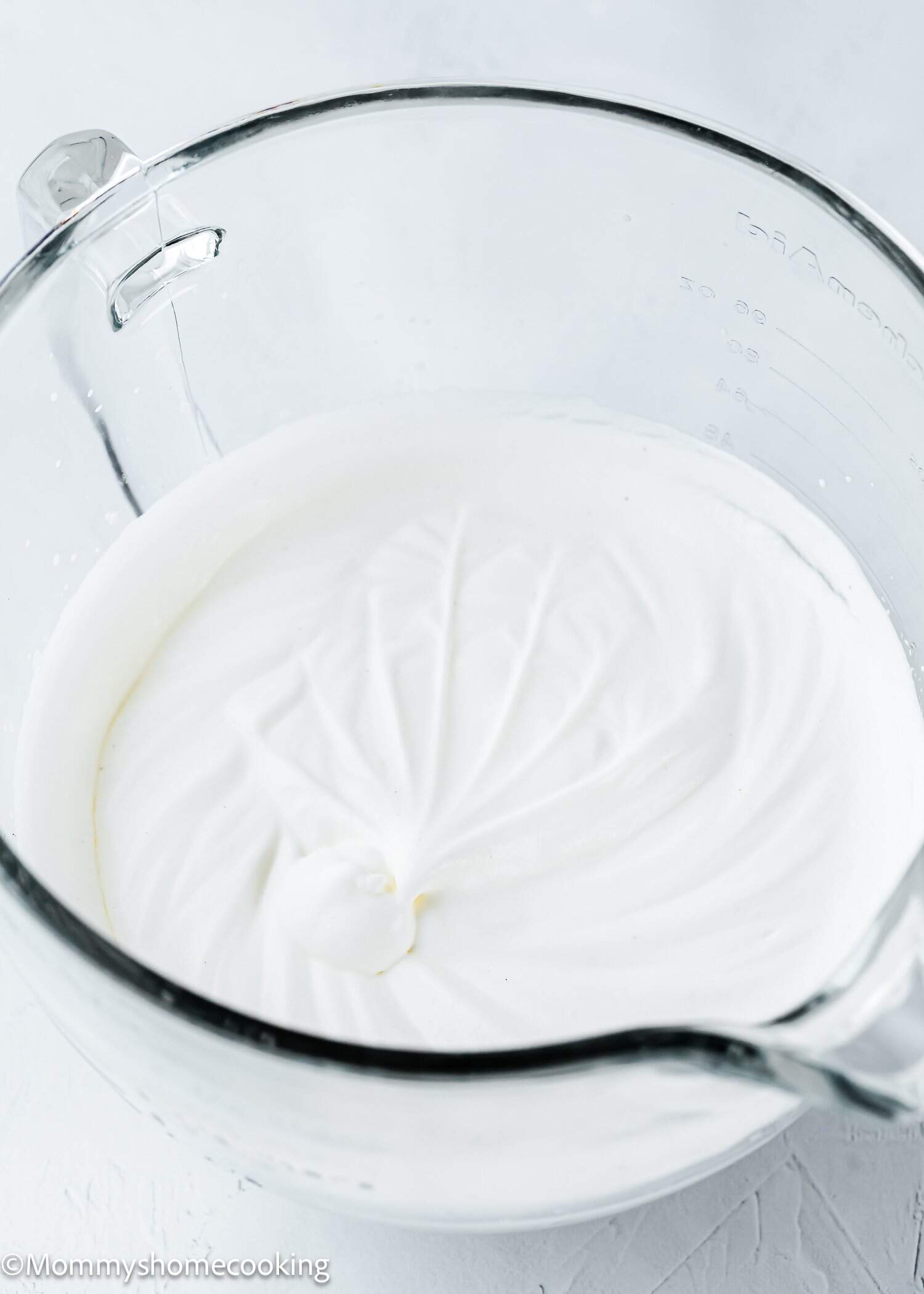 A glass mixing bowl filled with whipped, glossy white meringue on a light surface.