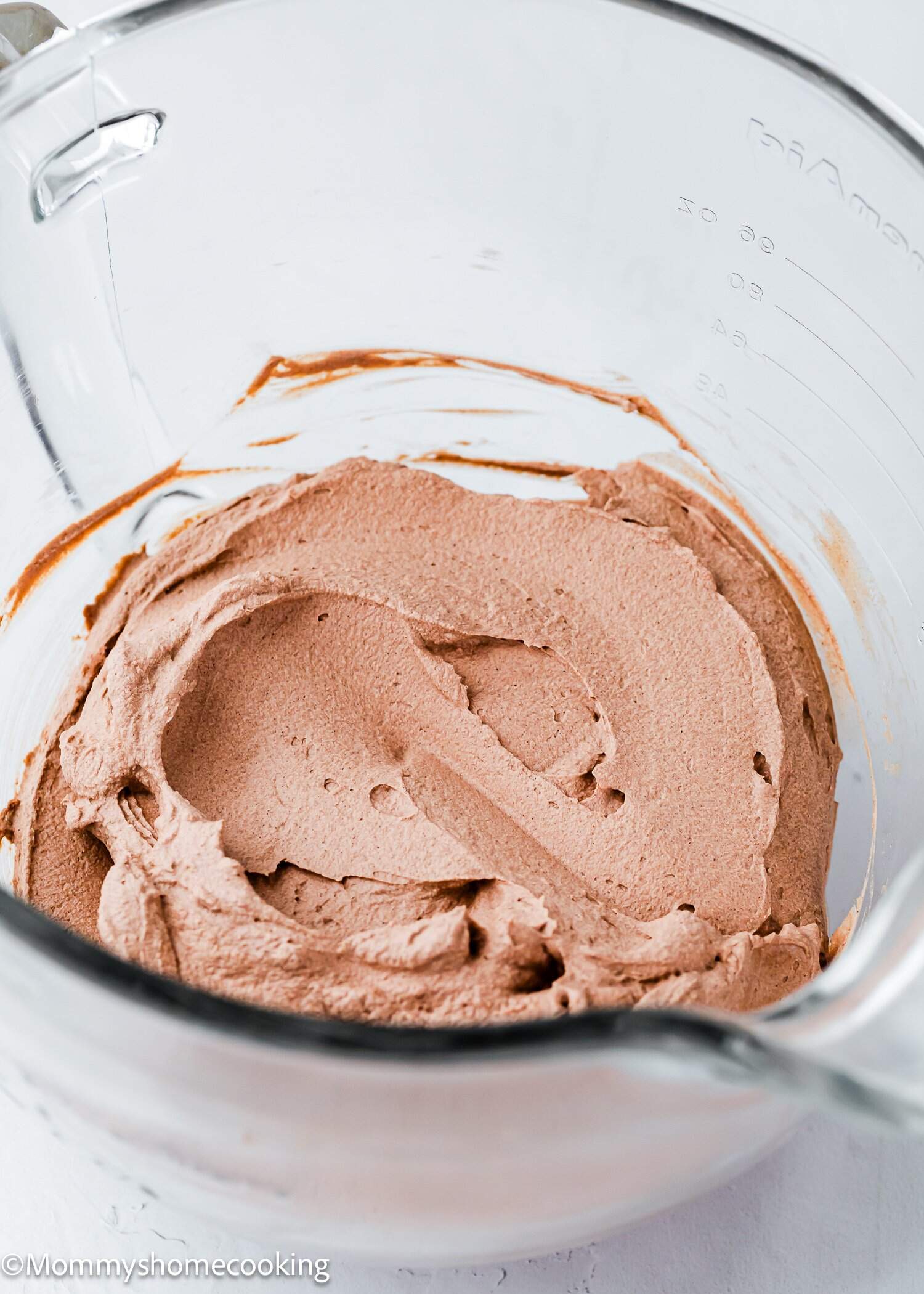 A glass mixing bowl filled with whipped chocolate frosting, with mixing marks visible on the surface.