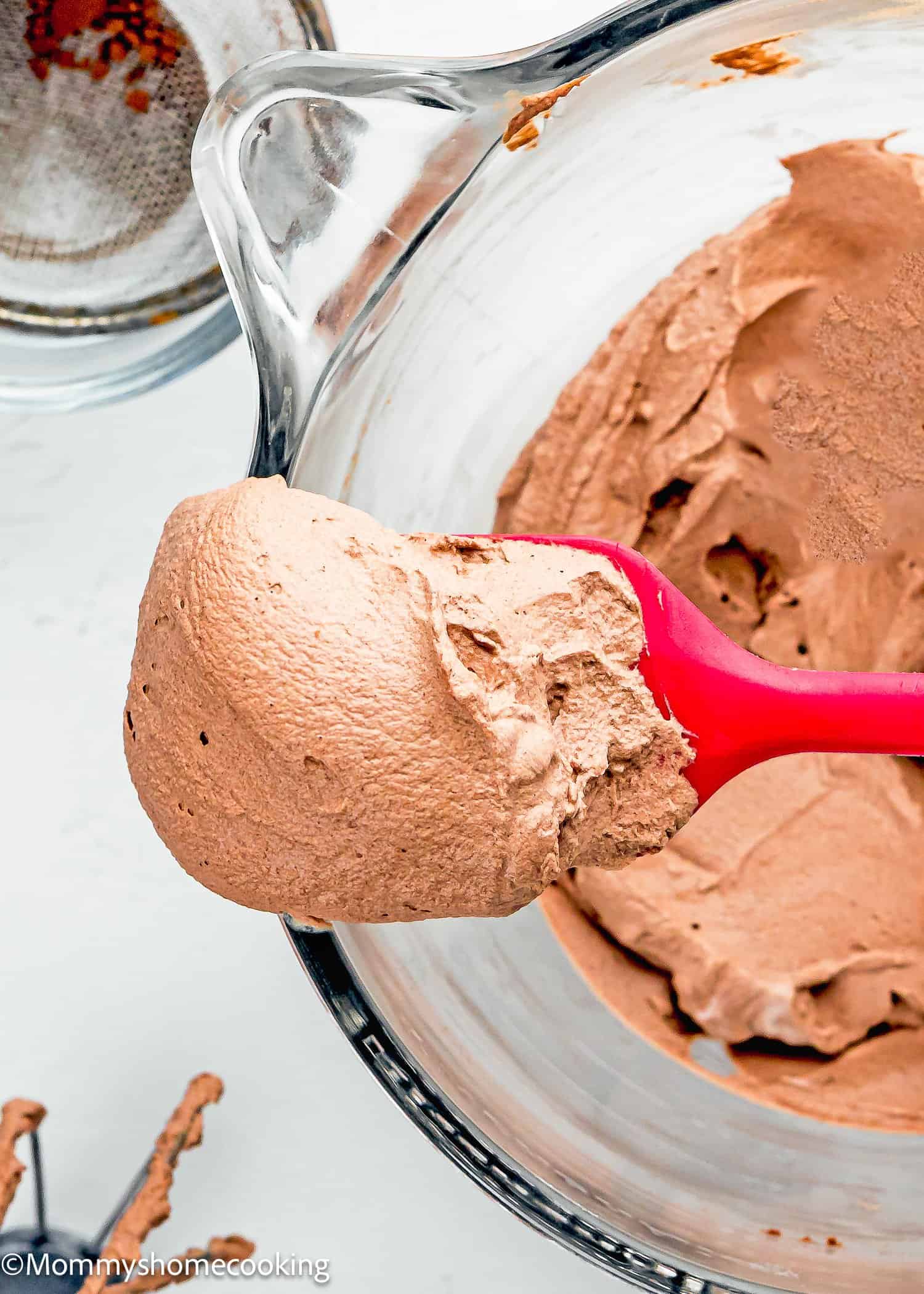 A red spatula holds a scoop of Chocolate Whipped Cream Frosting above a glass mixing bowl filled with more chocolate mousse.