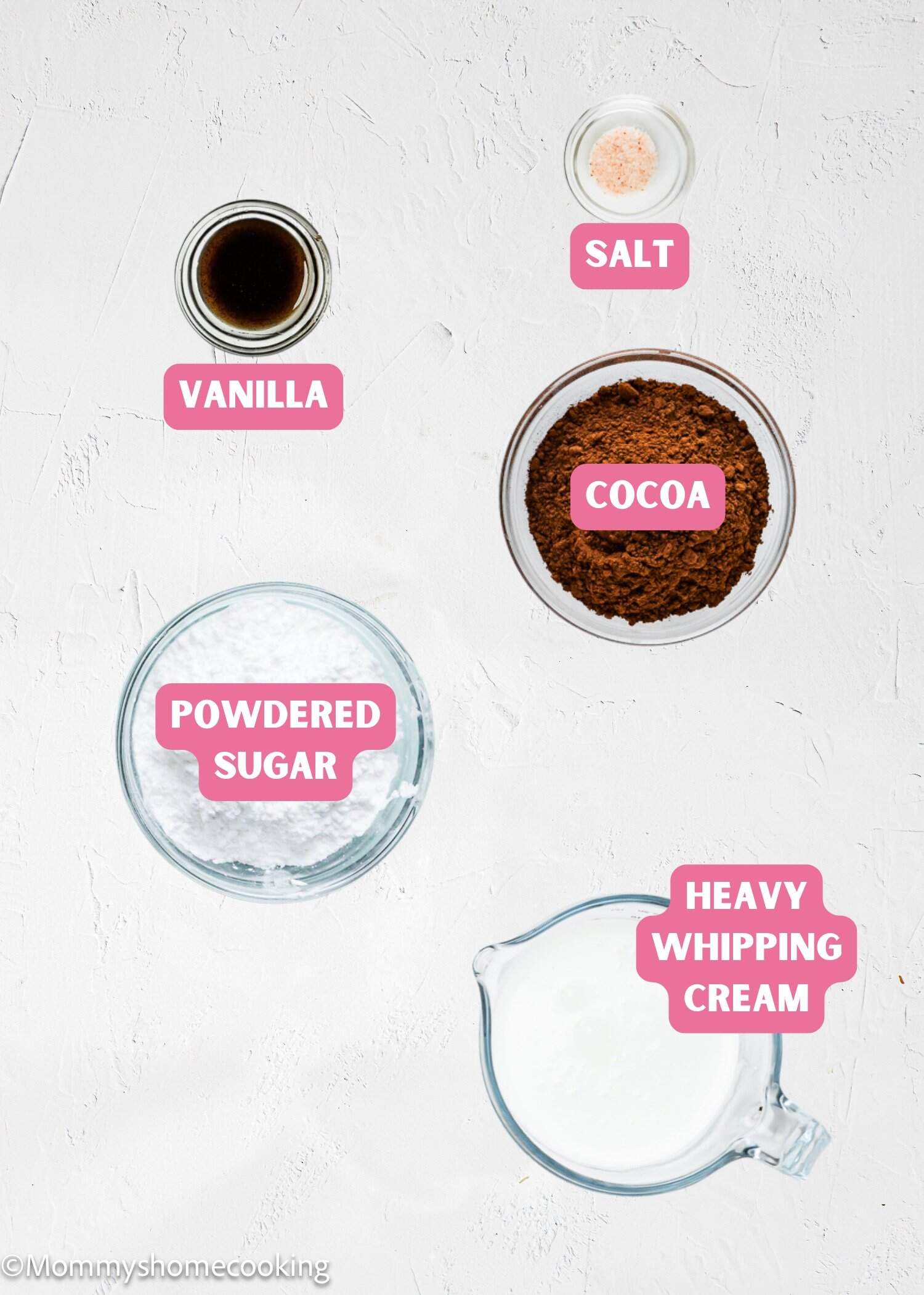 Five glass bowls containing vanilla, salt, cocoa powder, powdered sugar, and heavy whipping cream are arranged on a white surface, each labeled with pink tags.