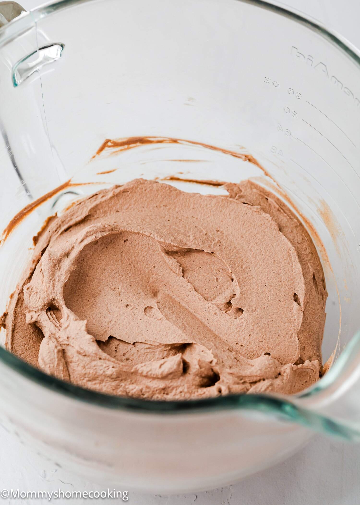 A glass mixing bowl containing whipped chocolate frosting, with streaks visible along the sides of the bowl.