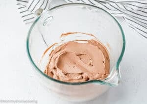 A glass mixing bowl containing chocolate whipped cream sits on a white surface with a striped cloth in the background, perfect for topping an Eggless Chocolate Pudding Cake.