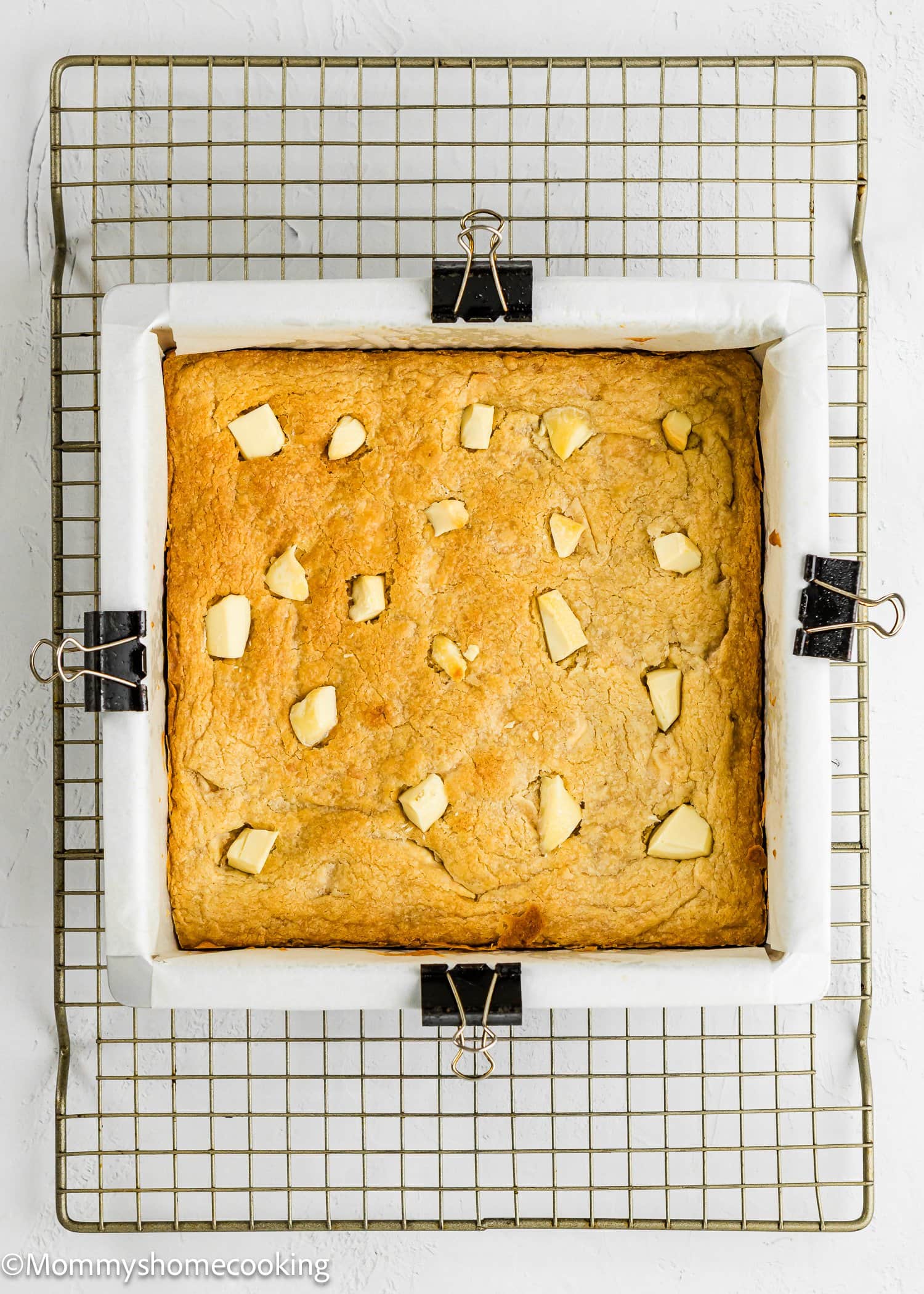 Square cake with chunks of butter on top, baked in a parchment-lined pan, cooling on a wire rack with binder clips holding parchment in place.